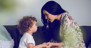 A black-haired mother having a tough conversation with her curly-haired toddler while sitting on a b...