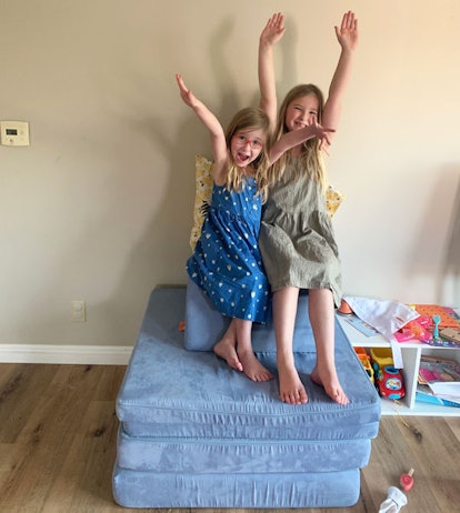 Two blonde girls sitting on a blue mattress, smiling and raising their arms in the air.