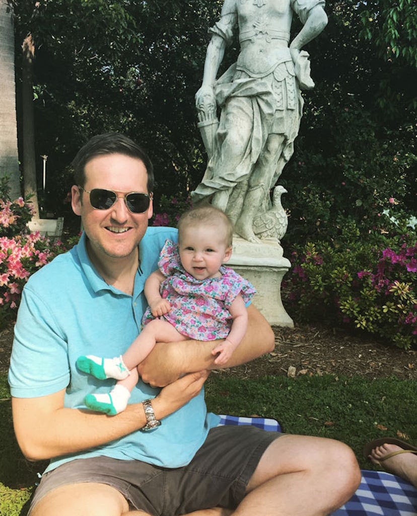 A father holding his smiling daughter in a pink dress in his hands with a statue behind them