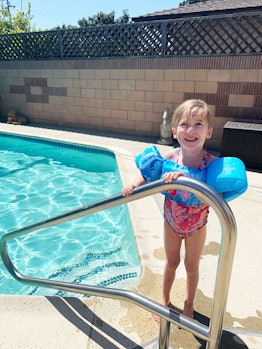 A blonde girl wearing a pink one piece swimming suit and smiling next to a pool