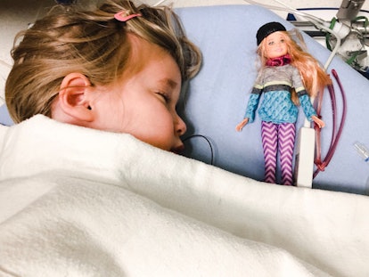 A blonde girl sleeping, covered with a white blanket with her doll beside her in a hospital bed