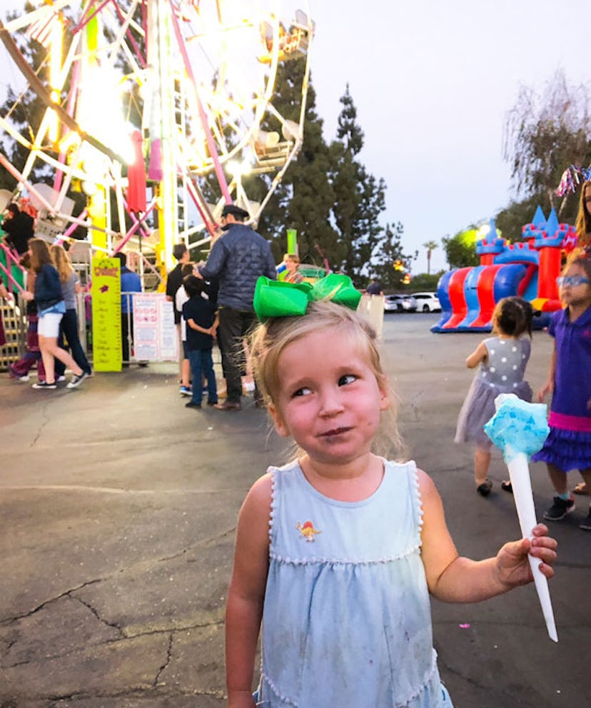 A blonde-haired girl wearing a blue dress, eating blue cotton candy in an amusement park