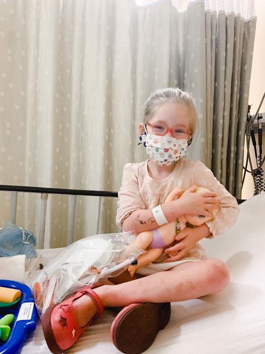 A blonde girl wearing pink glasses and a mask while sitting on a hospital bed and holding her doll