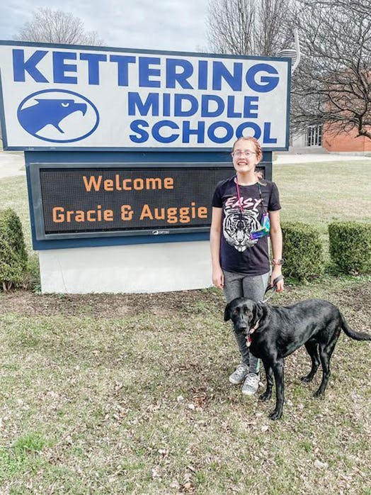 Gracie and her service dog Auggie in front of a "Kettering Middle School" sign and "Welcome Gracie a...