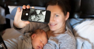 A mom lying in bed with her newborn on her chest, holding a phone up and taking a selfie