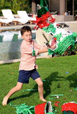 A boy in an orange shirt and navy-blue shorts hitting a pinata in a backyard during a family's no-oc...