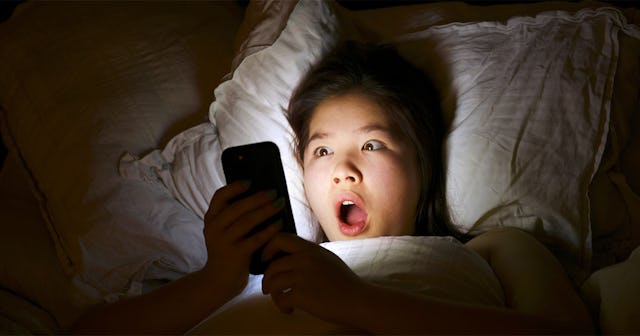 A woman with a surprised facial expression lying in bed and looking at her phone in a dark room