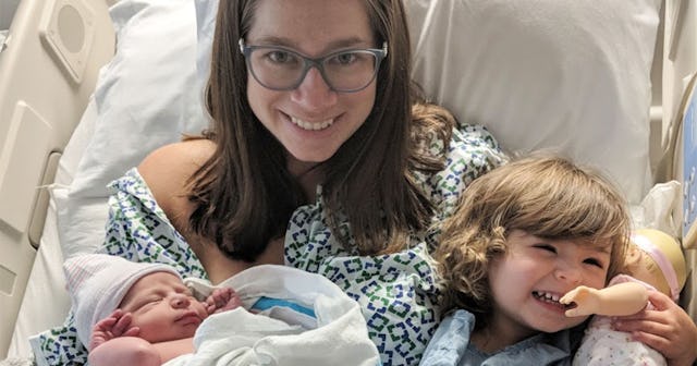 A woman after giving birth to her baby, holds it in one arm while her other, older child is sitting ...
