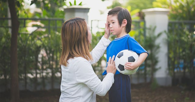 A woman in a white shirt looking at her son and leaning her hand on his face, while he holds a footb...