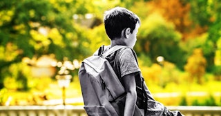 A small boy sitting alone with a school backpack on his back during summer