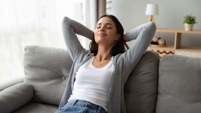 A relaxed woman sits on a gray sofa in a white tank top, gray sweater, and jeans, treating herself l...