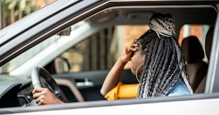 A woman with brown and blonde small box braids sitting in her car with one hand on the wheel and the...