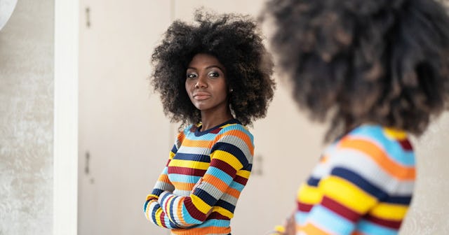 Black haired woman with curly hair in blue, orange, grey and yellow sweater looking in a mirror