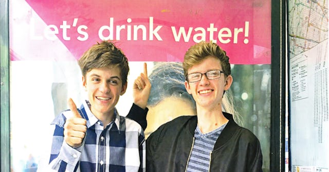 Two boys, one showing a thumbs up and another one pointing at a sign saying, "Let's drink water!"