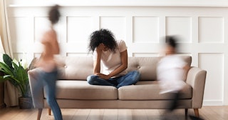 A mother sitting on a couch with her head down with her son and daughter playing next to her blurred...