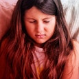 A nine-year-old girl with long brown hair feeling uncomfortable while laying on the bed with white s...