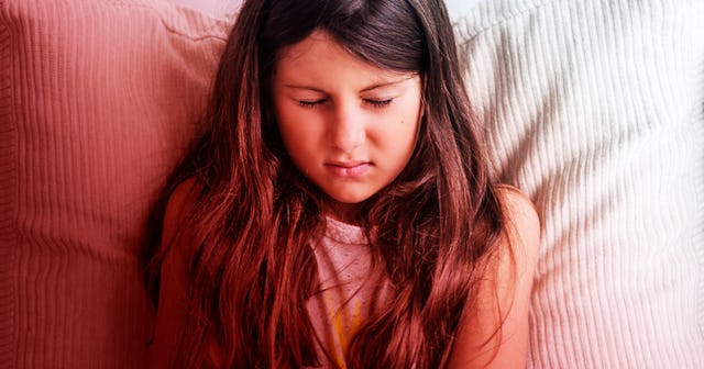 A nine-year-old girl with long brown hair looking uncomfortable while lying on the bed with white sh...