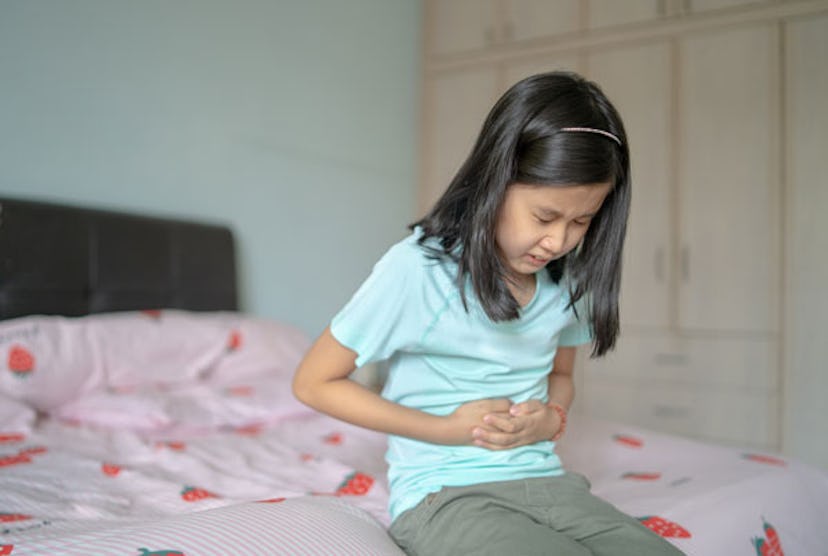 A nine-year-old girl with long dark hair sitting on the bed while uncomfortably holding her stomach.