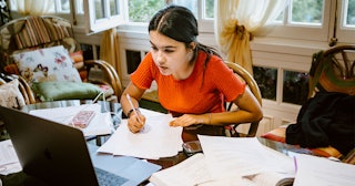 Brunette tween girl in an orange shirt writing in her notebook looking into her laptop during online...