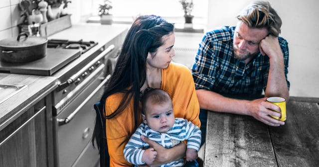 Worried parents looking at each other while sitting at the kitchen desk, with their baby in mother's...