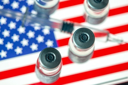 A syringe next to four glass vials with vaccine on top of a blurred American flag 