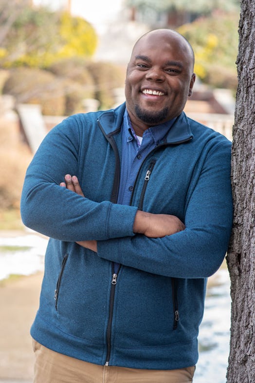 Author Kwabe Mbalia widely smiling while wearing a blue sweater and light brown pants, leaning on a ...