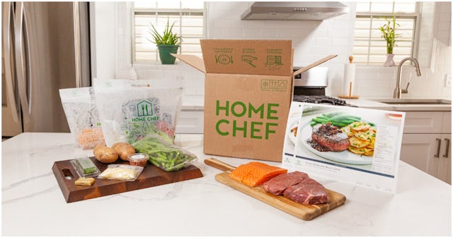 A Home Chef box next to two wooden platters with veggies, meat and fish on a whit marble kitchen cou...