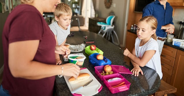 Mother wearing a red shirt packing school lunch for her kids, who are waiting patiently with the fat...