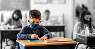 A classroom with children sitting and writing in their notebooks while wearing face masks instead of...