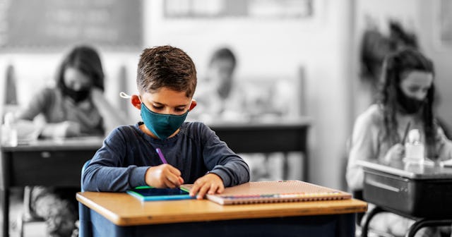 A classroom with children sitting and writing in their notebooks while wearing face masks instead of...