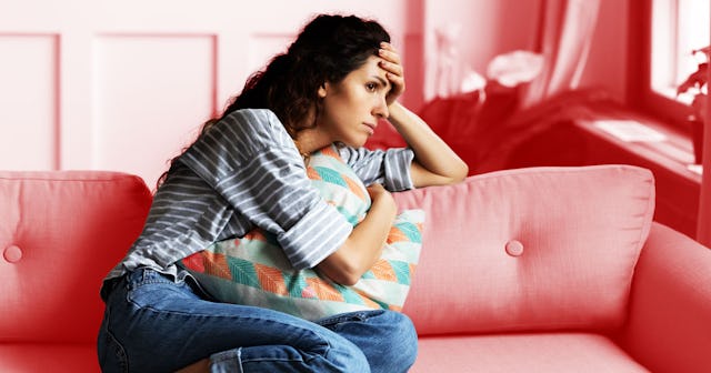 A worried mom sitting on a pink sofa, one hand holding a cushion and the other on her forehead while...