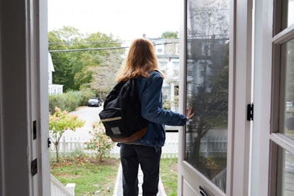 A girl in a denim jacket, gray pants, and a black and brown backpack is heading out of a house