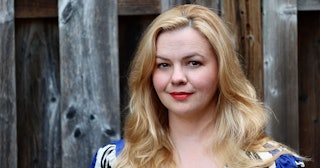 A portrait of Amber Tamblyn wearing red lipstick, smiling and looking straight in front of a wooden ...