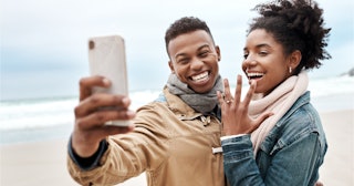 A couple taking a selfie and smiling while she’s showing off the engagement ring under $500 on her f...