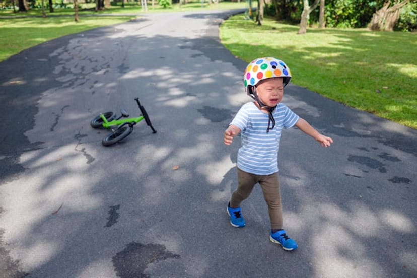 A crying kid with a helmet on walking away during a tantrum from a green bike on the street in the b...