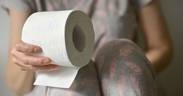 A woman sitting curled up due to her urinary retention while holding a roll of toilet paper