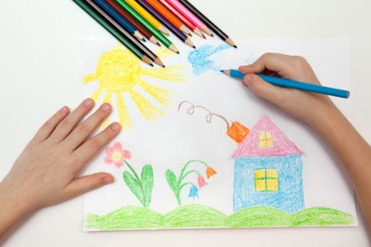A kid drawing a house with wooden crayons on a sheet of a paper