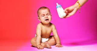 A baby sitting and crying while someone handles it a bottle.
