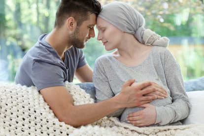 A pregnant woman who has cancer in a grey shirt and headscarf is leaning against her husband's head