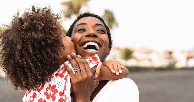 A woman with black hair smiling while her youngest spoiled kid kisses her