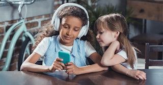 Two girls sat at a table, one has headphones on. Both are looking at Zigazoo - TikTok for kids on an...