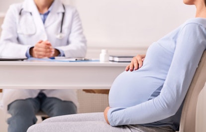 A woman is sitting in a doctor's office while holding her pregnant stomach and talking to a doctor a...