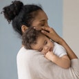 A dark-haired woman with her hair in a bun in a beige sweater holding her baby who had the vaccine r...