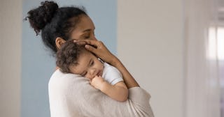 A dark-haired woman with her hair in a bun in a beige sweater holding her baby who had a legit vacci...