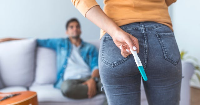 A woman hiding the pregnancy test behind her back while her husband is sitting on the couch