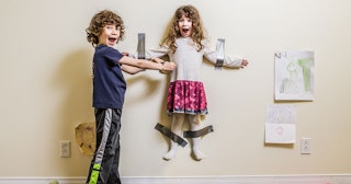 A brother and a sister laughing while the girl is hanging from the wall, her arms and legs taped by ...