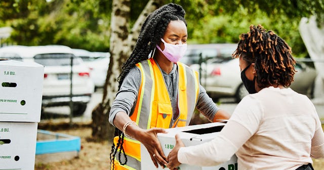 A woman in an orange vest with dreadlocks giving another woman with curly hair a white box