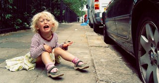 Little girl with autism sitting on a sidewalk next to a parked black car and crying hysterically.