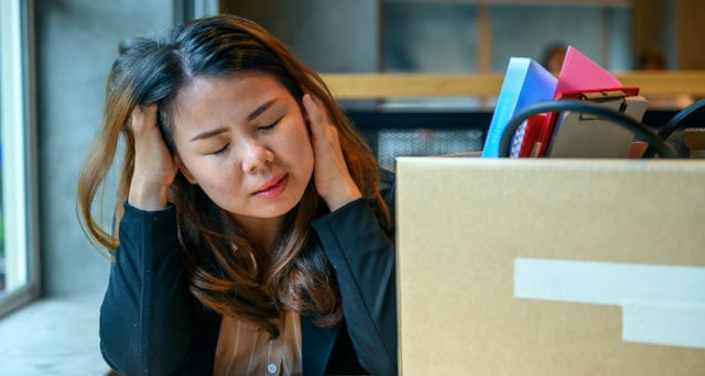 An exhausted-looking working mom, holding her neck and hair, is sitting in front of a box of office ...