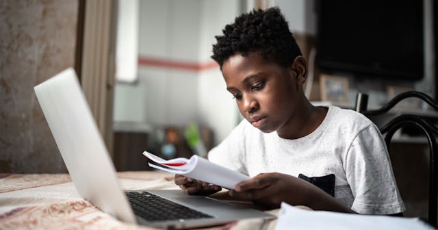 Gifted child sitting at a table with a laptop, reading from a notebook